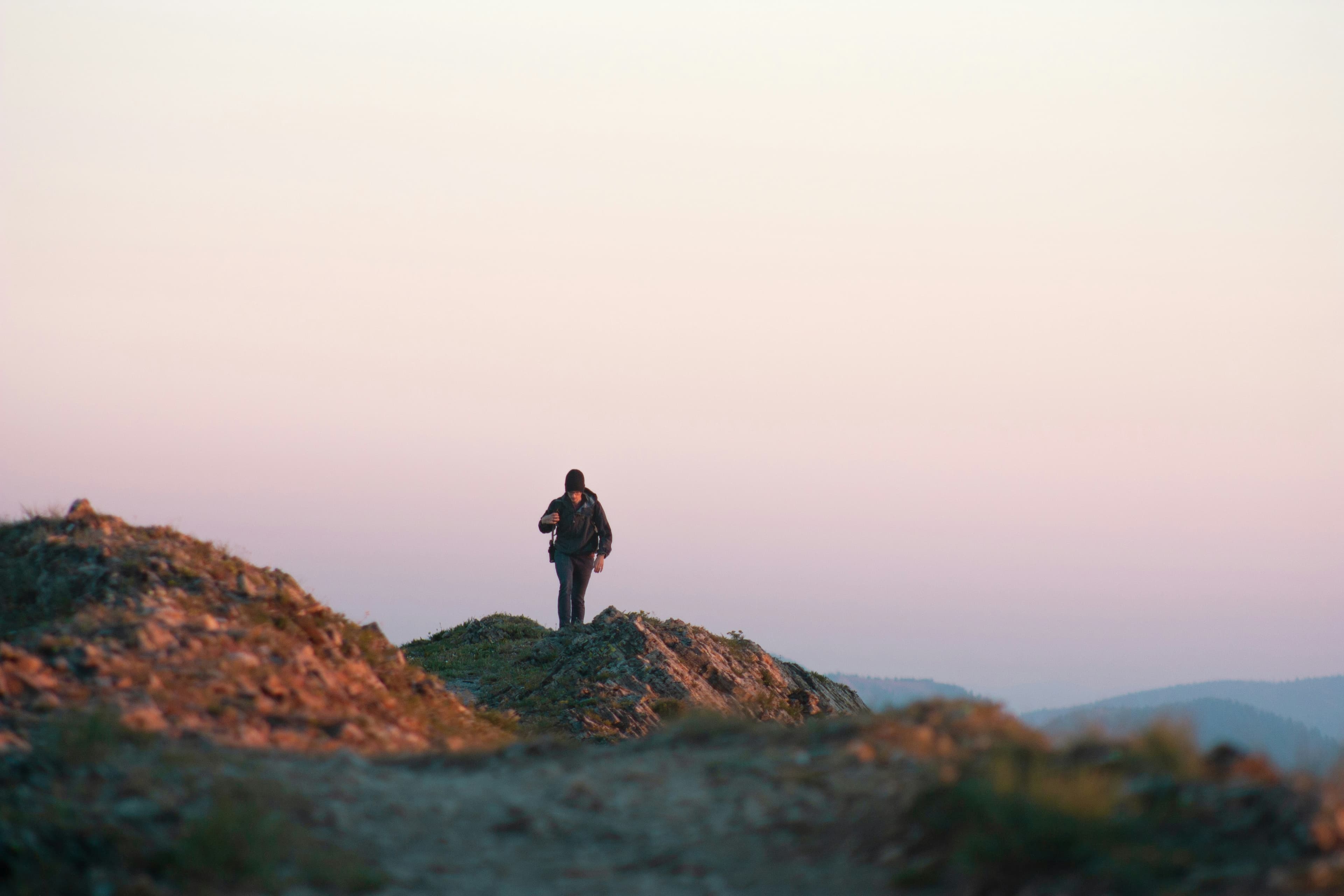 A lone figure on a rocky outcrop at sunrise, soft pastel sky filling the frame.